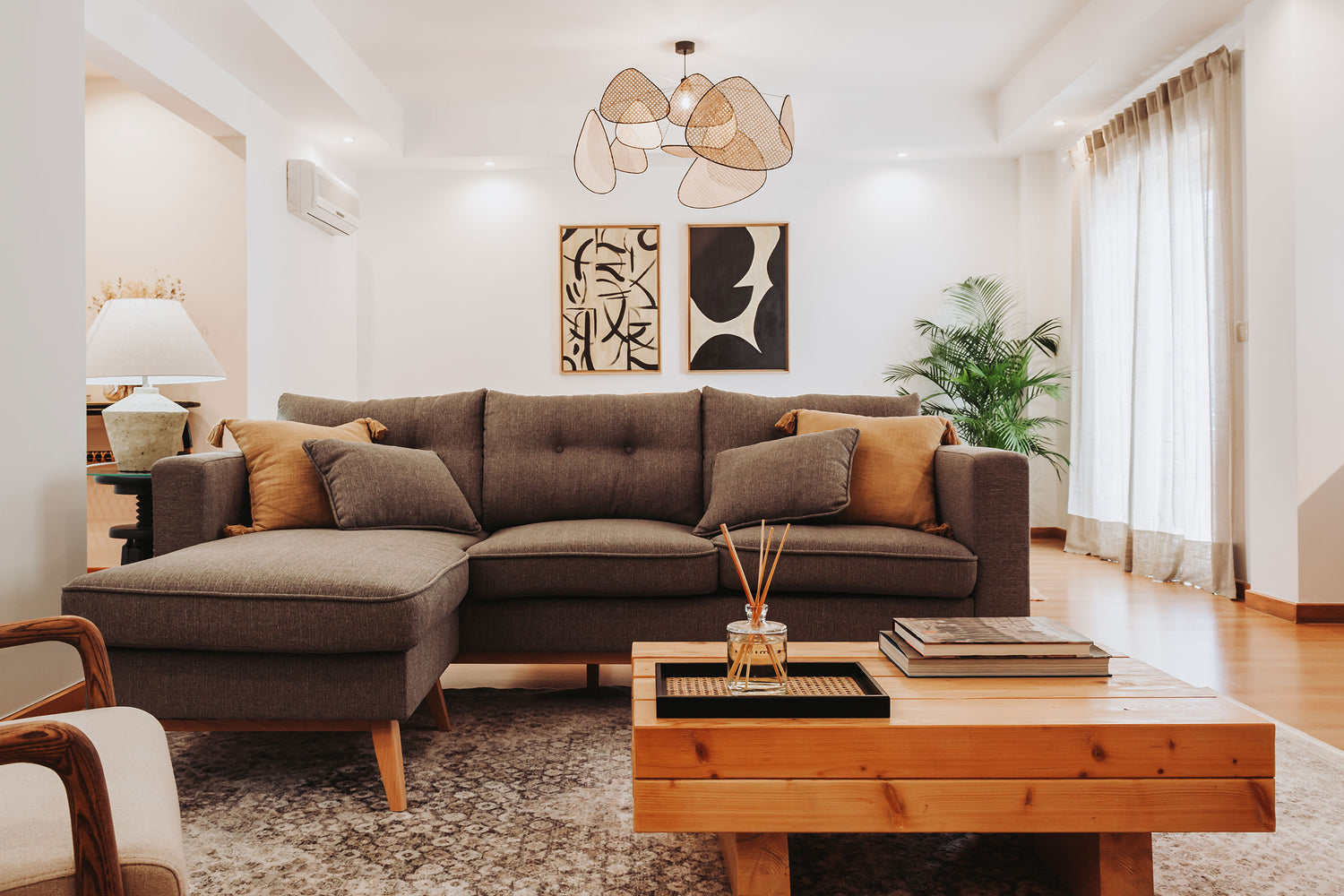 Sala de estar renovada, com um sofá cinzento com chaise longue, uma mesa de apoio de madeira e dois quadros com formas orgânicas de fundo.
